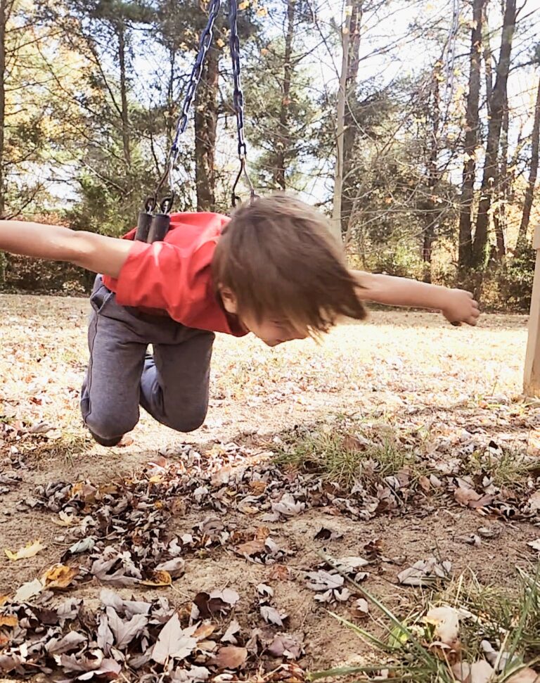 Boy in red shirt happily swinging outside. Leaves on the ground.